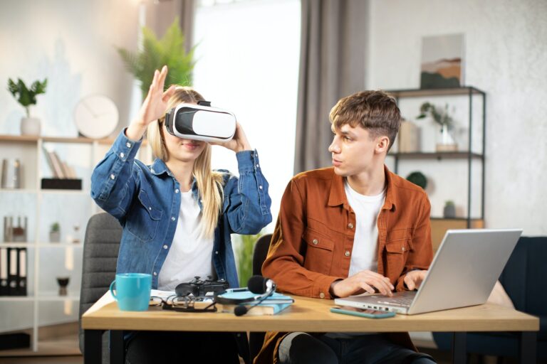 Happy young friends sitting at the table and using modern gadgets for work or study